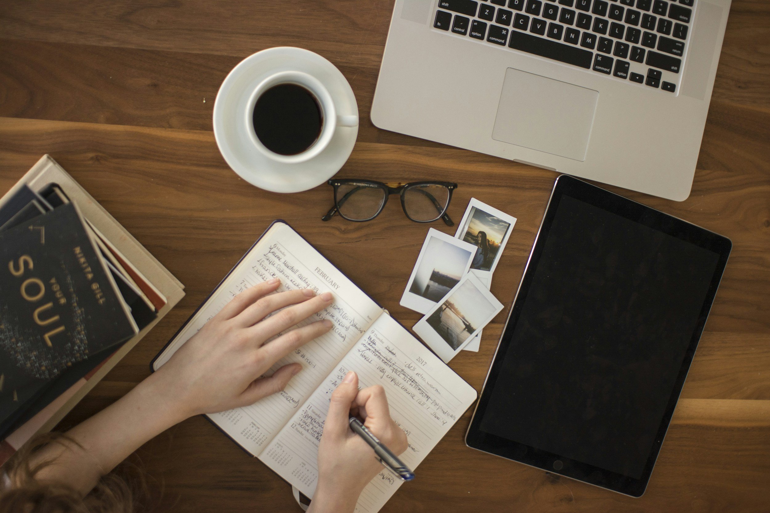 Scrivania con portatile, tablet, caffè e un'agenda su cui una mano prende appunti